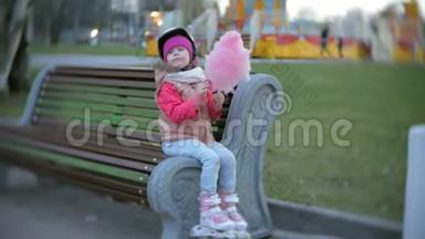 美丽的小<strong>女孩</strong>坐在游乐园的长凳上，吃着滚<strong>滑轮滑</strong>冰鞋上滚来滚去的粉红色甜棉糖